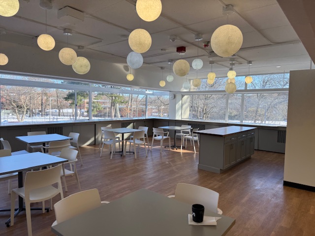 A bright common room at SAO with globe pendant lights, tables, and big windows onto the wooded campus.